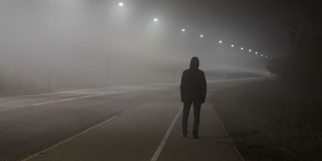 A lone figure standing on a foggy road at night, capturing the unease behind why do I feel like I’m being watched.